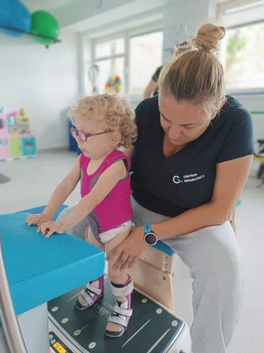 Terapeutka wspiera dziewczynkę z SMA podczas ćwiczeń pionizacji i obciążania kończyn dolnych na platformie rehabilitacyjnej. 