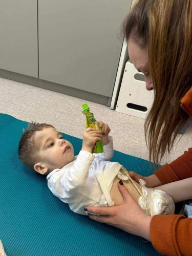 Małe dziecko leży na macie rehabilitacyjnej i sięga po zabawkę, wykonując ćwiczenia wspierające aktywność ruchową oraz koordynację wzrokowo-ruchową. 