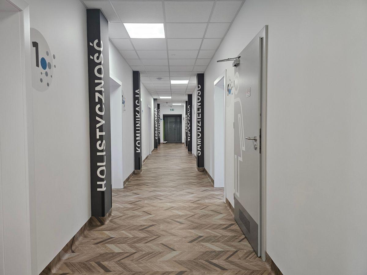 Jasny korytarz w Centrum Grawitacja prowadzący do gabinetów terapeutycznych i sal szkoleniowych, oznaczonych nazwami specjalistycznych obszarów terapii. 