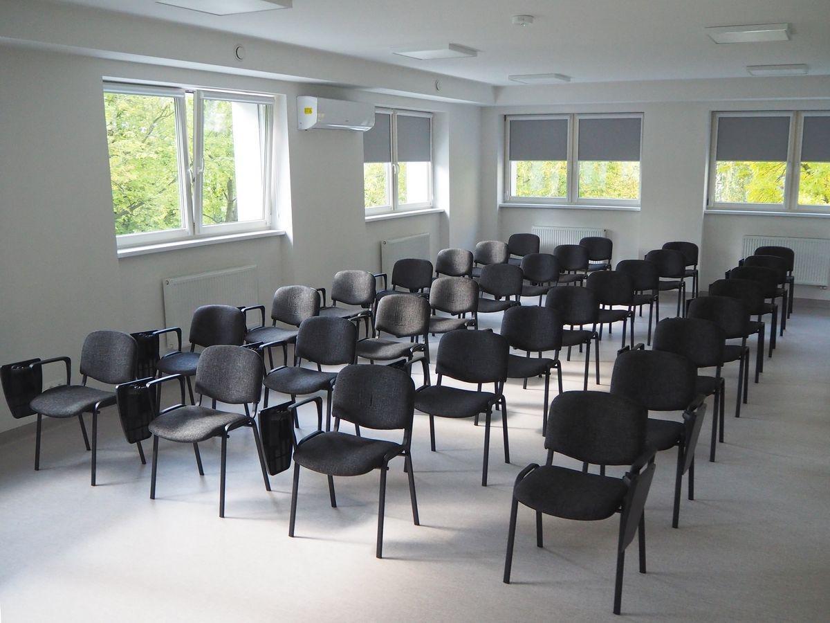 Jasna sala konferencyjna z miejscami siedzącymi dla uczestników oraz dużymi oknami zapewniającymi naturalne oświetlenie. 